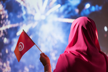 Muslim woman in a scarf holding flag of Tunisia during fireworks at night.