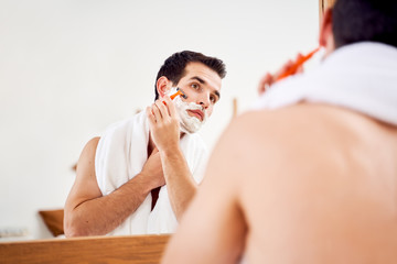 Young male with foam on his beard shaves near mirror in morning
