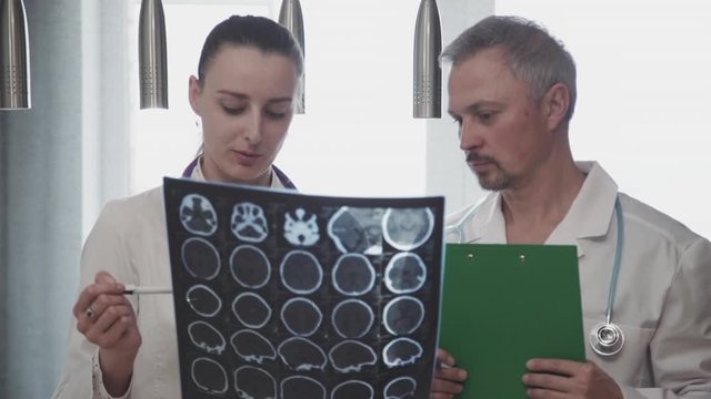Man doctor and woman talking while looking at x-ray picture in modern clinic. Front view of young colleague having conversation about work, holding MRI result in hand. Two experts wearing white coats