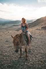 a young girl with red hair sees riding a red horse at sunset along a hill scorched by the sun against a background of high snow peaks - 316955304