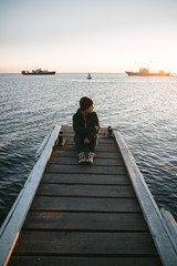young traveling girl sits on a pier on the background of the sea and looks at the sunset and ships  - 316958130