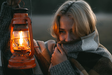 close portrait of a young girl with blond hair in a warm woolen sweater wrapped in a plaid, she is standing in a field at dawn with a kerasin lamp in her hands - 316958597