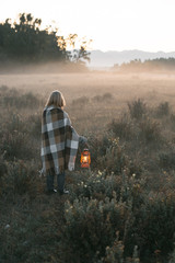 vertical shot of a young girl with blond hair in a warm woolen sweater wrapped in a plaid, she is standing in a field with tall dry grass at dawn with a kerasin lamp in her hands - 316958774