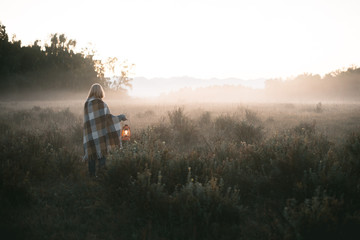 young girl with blond hair in a warm woolen sweater wrapped in a plaid, she is standing in a field with tall dry grass at dawn with a kerasin lamp in her hands - 316959142