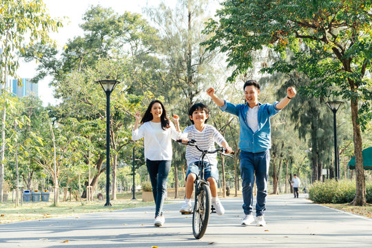 Happy Asian father and mother teach their son to ride a bicycle, Cheerful parent raise hands up in the air to support kid encouragement, family do activity together at park concept