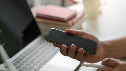 Cropped shot of businessman holding  smartphone  in modern workspace with laptop and office supplies