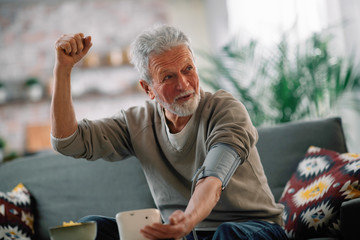 Old man sitting in living room. Happy senior man checking blood pressure. 