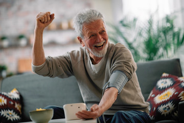 Old man sitting in living room. Happy senior man checking blood pressure. 