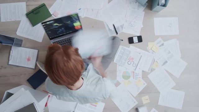 Time lapse top shot of Caucasian man with red hair sitting on floor, working on laptop with data graphs running on screen, analyzing charts, studying and writing on documents