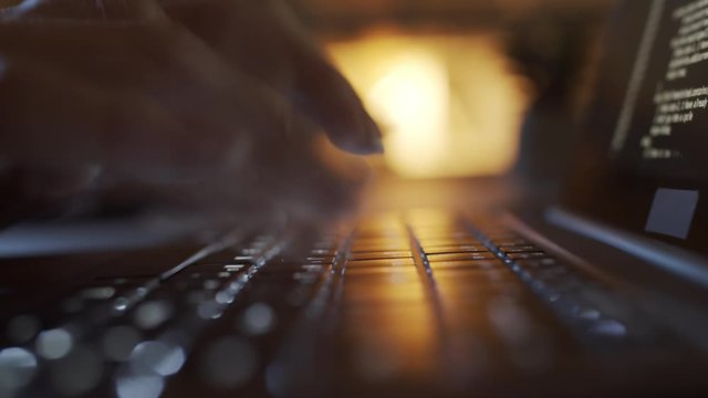 Fast motion close-up shot of laptop, hands and fingers of unrecognizable female programmer typing on keyboard late at night, illuminated by lamp in background, and lines of symbols running on screen