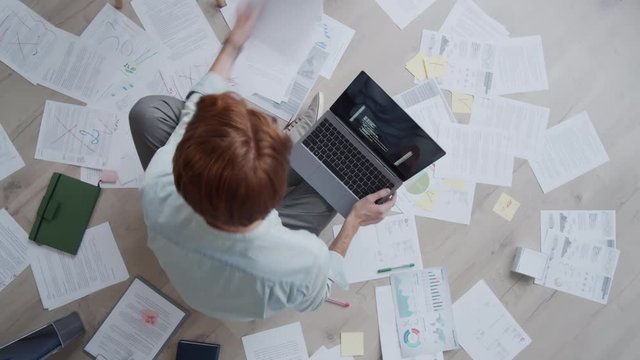 Time-lapse top shot of busy Caucasian man with red hair sitting on floor at home, writing computer code on laptop and working with documents, charts and diagrams laid out around him