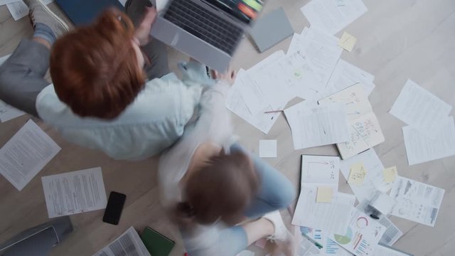 Timelapse top shot of man sitting on floor at home, typing and working on graphs and diagrams on laptop, while woman is writing in notebook and on clipboard, studying documents and charts