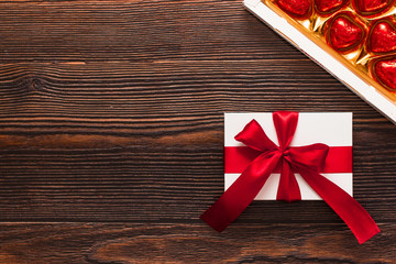 White present with a red ribbon and a box of chocolate red hearts isolated on a dark wooden background. Top side view of a celebrating warm flatlay. Valentines day and Christmas concept. Copyspace.