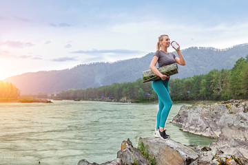 Blond beautiful strong girl standing on the stone cliff ground resting after sport outdoors drink...