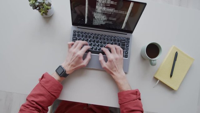 Timelapse rotating top shot of hands of busy male programmer sitting and working at desk, writing computer code on laptop, drinking coffee from mug, and lines of symbols running on screen