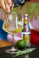 Alcoholic cocktail gin and tonic with lime, rosemary, dried fruit, cucumber on a wooden table, barman's hand with bar tongs spreads decorations..