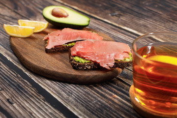 Avocado toast with whole wheat bread, guacamole and smoked salmon on cutting board. Near slices of lemon and half an avocado. Healthy breakfast concept. Wooden background, selective focus. 