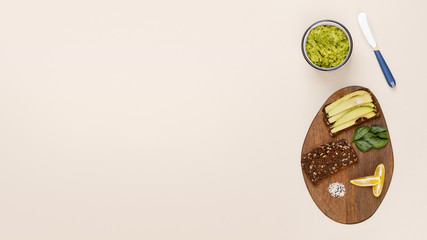 Whole wheat toast with avocado slices, fresh basil leaves, lemon slices and sesame seeds on wooden board. Near avocado paste and guacamole in bowl and knife for spreading. Flat lay, beige background.