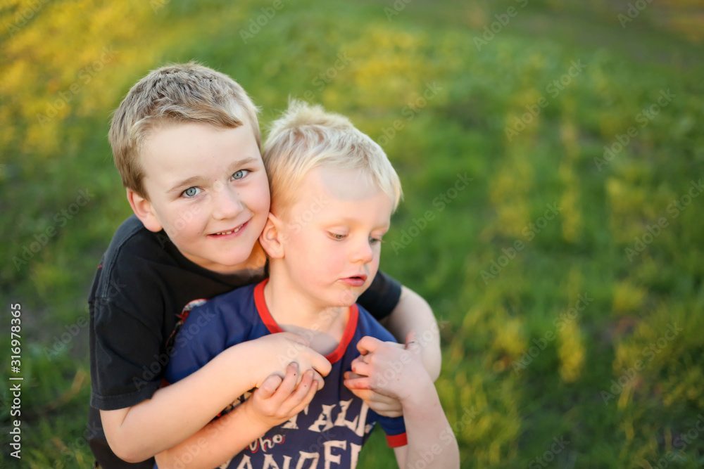 Wall mural Two young brothers hugging with faces pressed together. Little boy with missing teeth. Children playing at golden hour.