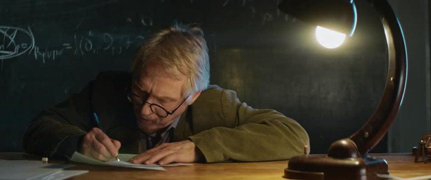 Aged Professor in glasses working, writing smth on paper sheet. desk lamp light. late night, thinking cogitative process, origin of idea, smart wise man