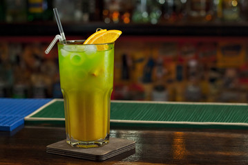 Green cocktail on bar counter. Drink with martini, gin, rum, tequila and fruit on the background of bottles with alcohol. Glass with booze on a wooden board.