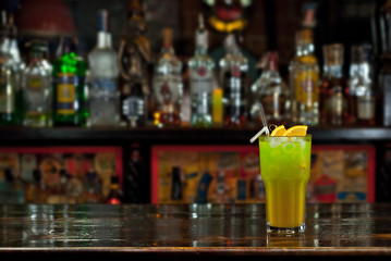Green cocktail on bar counter. Drink with martini, gin, rum, tequila and fruit on the background of bottles with alcohol. Glass with booze on a wooden board.