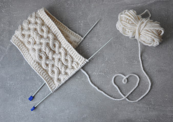 Knitted white scarf with ornaments and knitting needles on a gray background. The heart of the threads on the concrete texture
