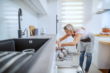 Obraz premium Worthy caucasian blond housewife in apron standing in domestic kitchen and putting dishes in dishwasher.