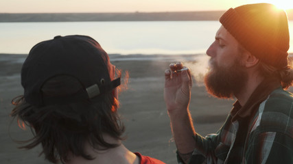 Two friends smoking joint with weed at seashore at sunset