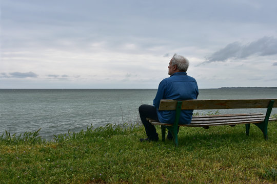 Senior man sitting on an old wooden bench above the see in a cloudy day, looking to the horizon.  Nostalgic mood. 