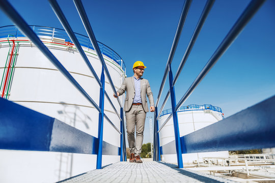 Young handsome caucasian businessman in suit and with helmet on head walking on bridge and looking at his refinery. In background is tank with oil.