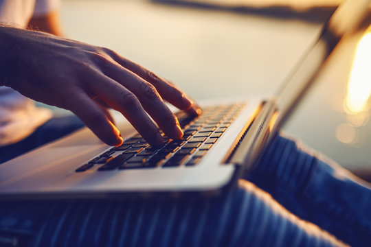 Cropped picture of Caucasian man sitting in nature and using laptop. In background is river and sunset. Selective focus on hands.