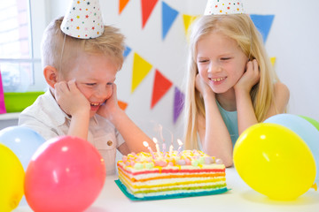 Fototapeta na wymiar Two blonde caucasian kids boy and girl looking at birthday rainbow cake with burning candles and making wishes at birthday party. Colorful background.
