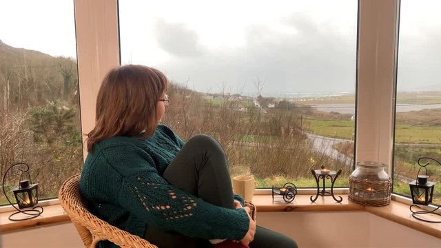 woman sits in a cozy wicker chair on a light closed veranda in the mountains and smiles, clutching her legs in warm high socks with fur, concept chillin and relaxation