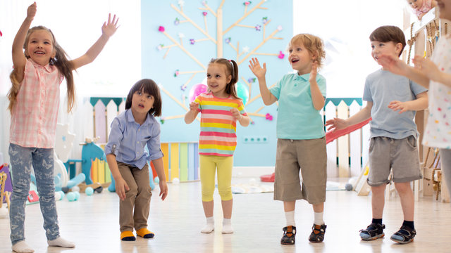 Cheerful kids stand semicircle on floor in kindergarten or daycare centre. Preschoolers have fun indoors, playing games