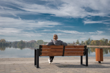 A young guy sits on a bench by the lake. A warm calm day, light and white clouds in the sky