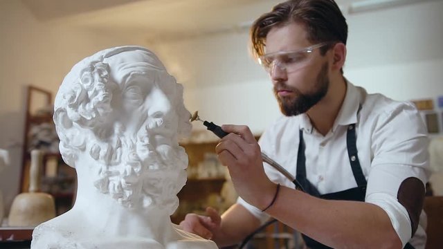 Slow motion of assured good-looking modern bearded sculptor which polishing the sculpture's head details in special glasses