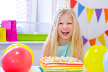 Obraz na płótnie Canvas Blonde caucasian girl laughing at camera near birthday rainbow cake. Festive colorful background with balloons.