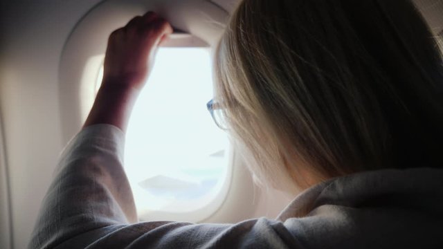A woman opens the porthole curtain and looks out the window of the plane.