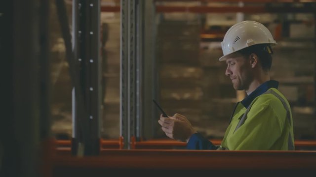 Male Industrial Worker in the Hard Hat Uses Mobile Phone While Walking Through Warehouse