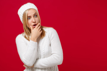 Shocked young woman in white sweater hat isolated on red wall background, studio portrait. Healthy fashion lifestyle, cold season concept. Mock up copy space. Looking aside, put hand prop up on chin.