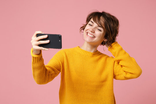 Smiling young brunette woman girl in yellow sweater posing isolated on pastel pink background. People lifestyle concept. Mock up copy space. Doing selfie shot on mobile phone, putting hand on head.