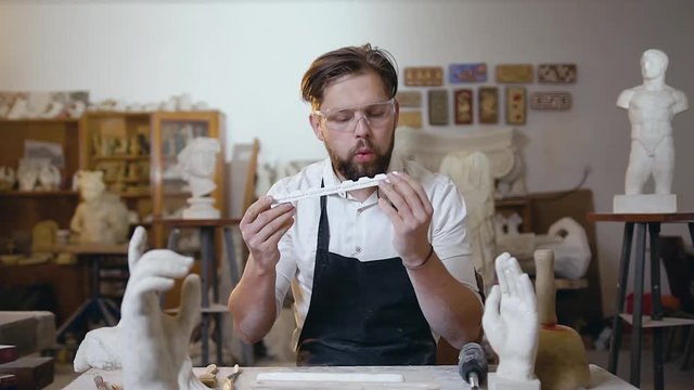 Studio interier where handsome modern bearded stonemason sitting at his workplace and looking at the detail from the limestone to the sculpture