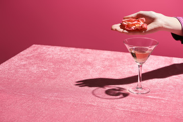 cropped view of woman holding rose petals above glass of rose wine on velour cloth isolated on pink, girlish concept
