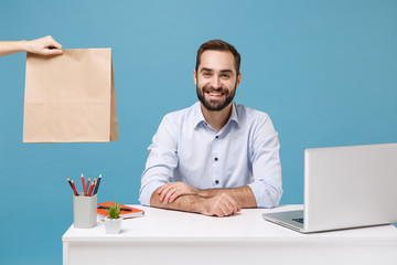 Smiling man in shirt sit work at desk with pc laptop isolated on blue background. Food products delivery courier service from shop or restaurant to office Take brown clear empty blank craft paper bag.