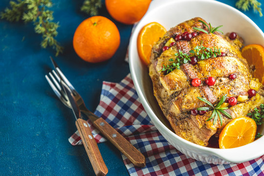 Roasted pork in white dish, christmas baked ham with cranberries, tangerines, thyme, rosemary, garlic on light table surface