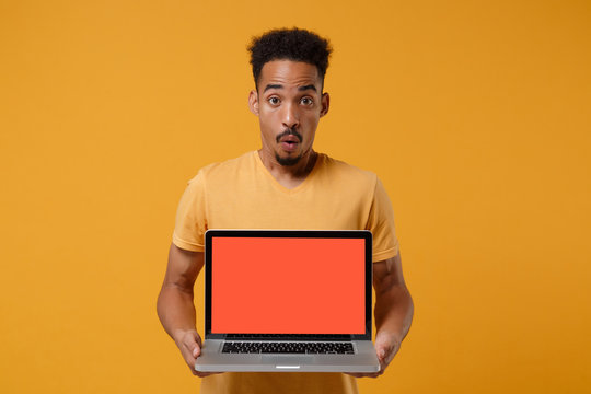 Shocked young african american guy in casual t-shirt posing isolated on yellow orange wall background. People lifestyle concept. Mock up copy space. Hold laptop pc computer with blank empty screen.