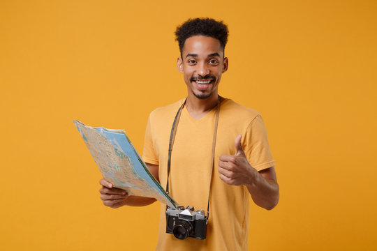 Smiling young african american guy in t-shirt with retro vintage photo camera posing isolated on yellow orange background. People lifestyle concept. Mock up copy space. Hold city map showing thumb up.