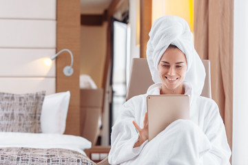 Entrepreneurial positive young business woman relaxing in spa hotel on business trip filling out an activity diary using a tablet while sitting on a couch in a bathrobe and a towel on her head