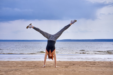 A middle-aged woman with red hair does gymnastics on the sandy shore of a large river. Cloudy spring morning.